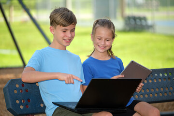 Two kids, teenager boy holding laptop computer and pretty young girl with digital tablet studying together outdoors sitting on bench. Education during quarantine concept