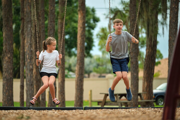 Two young teenage children, girl and boy playing on swings together outdoors on bright sunny...