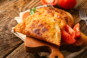 Cutting board with tasty chebureks and tomato slices on wooden background