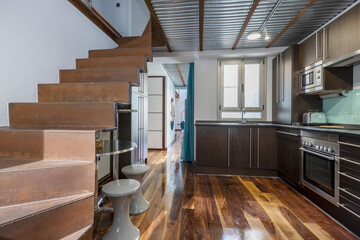 Modern kitchen of a loft apartment with dark wooden cabinets, windows, unpainted metal stairs and jatoba parquet flooring