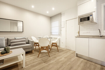 Studio apartment with an open kitchen with white cabinets and a round white dining table with resin chairs, a 3-seater sofa upholstered in gray fabric and a white wooden coffee table