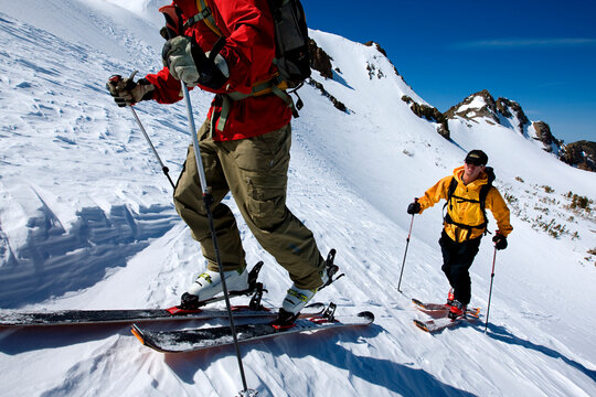 Two people skinning / backcountry skiing up a mountain on a blue sky day.