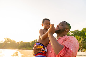 Happy African family on beach holiday vacation. Father playing and carrying little son at tropical beach at summer sunset. Dad with boy kid enjoy and fun outdoor lifestyle activity together at the sea