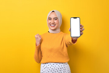 Beautiful excited Asian girl wearing hijab showing mobile phone with blank white screen, raising fist, celebrating good luck isolated on yellow background, mockup, copy space