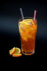 Orange juice in a tall glass with ice and straws on a black background, next to pieces of ripe citrus.