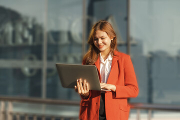 Obraz premium Using laptop, Smiling young Asia business woman leader entrepreneur in red color suit working outdoor urban street walk.