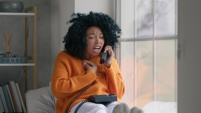 Upset girl with afro hairstyle got angry, sorting things out and hang up. Portrait of african american woman in orange sweater sitting at window with handset phone swear at boyfriend over the phone