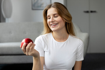 Happy beautiful woman with toothy smile looking at red apple sitting at home. Healthy food, vitamins and people concept 