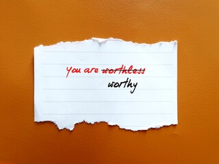 On orange background, torn paper with text YOU ARE WORTHLESS, crossed out to YOU ARE WORTHY - concept of overcome negative self talk, to have self respect and treat yourself with care