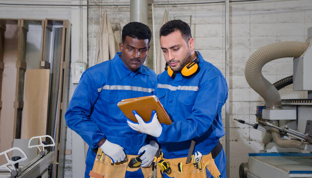 Young middle eastern labour in mechanic jumpsuit, ear muff and tool belt holding tablet computer, check out the production process while working in a furniture factory. A technician stand beside him.