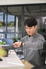 Handsome Asian male developer or office worker checking time on his smartwatch at his desk.