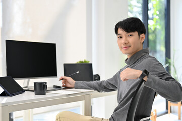 Handsome Asian male graphic designer or program developer sits at his desk in modern office