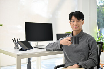 Asian male developer sits at his desk, holding a cup of coffee, smiling and looking at camera.
