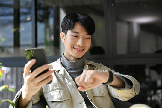 Smiling Asian man holding his phone, checking time on his smartwatch in the coffee shop.