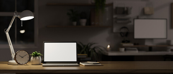 Laptop mockup, alarm clock, stationery, decor plant and table lamp on wood tabletop in dark office