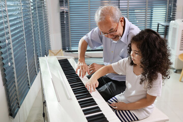 Fototapeta premium Happy smiling asian senior man sitting and playing piano while teaching grandchild in living room house indorrs. Musical and relaxation makes elder male happiness. Health care lifestyle concept.