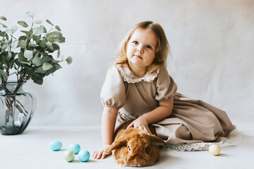 little blonde girl in vintage dress near retro background and vase with green branches eucalyptus gum tree, brown bunny rabbit spring day, happy childhood, simple and cozy life, happy Easter eggs hunt