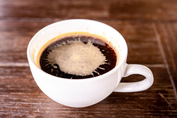 a steaming cup of hot black coffee on a wooden table. The coffee is dark and rich in color, with a thick layer of foam on top.