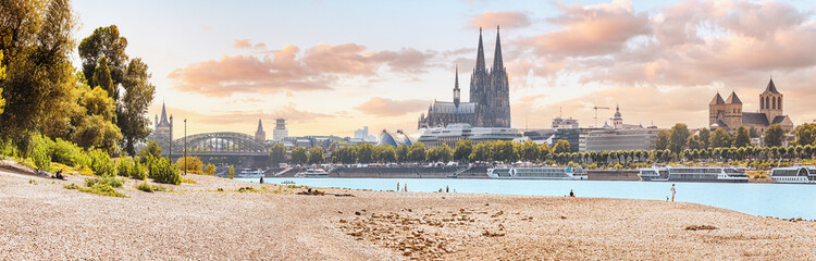 Panoramic view of the Rhine River beach and the Cologne skyline with recognizable architectural silhouettes of the famous Koln Cathedral - obrazy, fototapety, plakaty