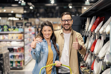A happy couple is standing next to the utensils and porcelain plates in supermarket and giving...