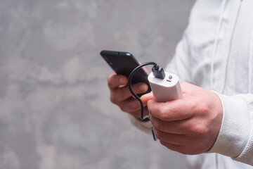Man holds a power bank in his hands and charges a smartphone against a concrete wall.