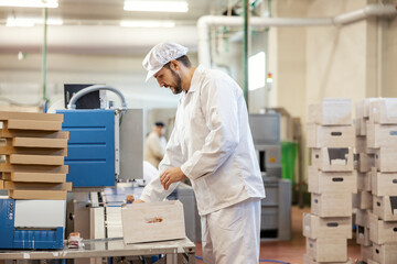 A meat industry worker is collecting meat products form conveyor belt and packing it into box.