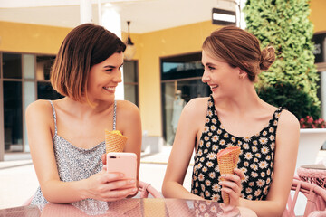 Two young beautiful smiling hipster women in trendy summer dress clothes. Sexy carefree models sitting in terrace cafe. Positive female eating tasty ice cream in waffles cone at sunny day