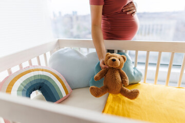 Pregnancy. Pregnant woman preparing nursery holding belly baby bump by crib holding teddy bear....