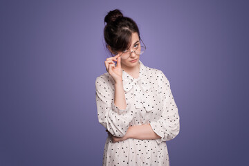 Portrait of serious suspicious young woman in eyeglasses looking at camera, posing on purple studio background.