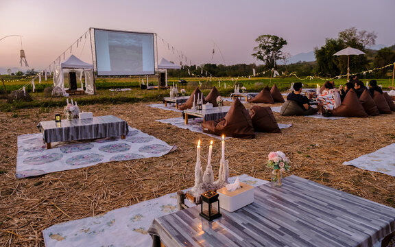 A Couple Of Men And Women Watching A Movie At An Outdoor Cinema In Northern Thailand Nan Province Out Over The Rice Paddies In Thailand, Green Rice Fields. 