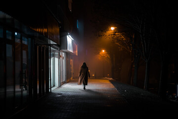 dark silhouette of a girl dressed in a long coat against the background of a night city with advertising showcases - 560013454
