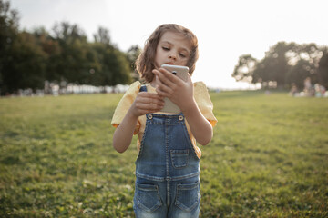 little girl 5 years old in yellow blouse and denim overalls looking to smartphone on sunny day in park