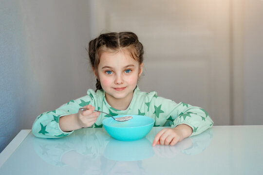 A cute little girl with an appetite eats porridge for morning breakfast in the kitchen. A hungry child holds a full tablespoon of delicious porridge experiencing pleasure delight and joy.