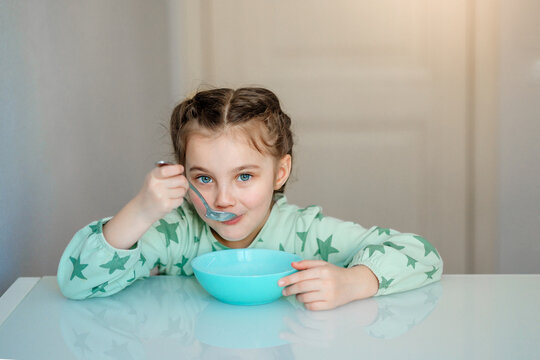 A cute little girl with an appetite eats porridge for morning breakfast in the kitchen. A hungry child holds a full tablespoon of delicious porridge experiencing pleasure delight and joy.