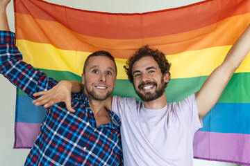 Two young gay lovers looking at each other affectionately. Two young male lovers standing together against a pride flag. Affectionate young gay couple sharing a romantic moment together.