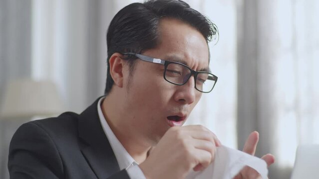 Close Up Of Sick Asian Businessman While Working With Laptop In The Living Room At Home. Sick Guy Blows Sneezes Wipes Snot Into Tissue
