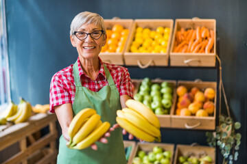 Mature woman works in fruits and vegetables shop. She is holding bananas.