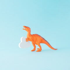 A creative aesthetic arrangement made of an orange dinosaur holding a white heart on a blue...