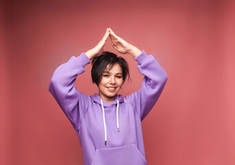 A happy young girl with short black hair makes a gesture of the roof of the house with her hands over her head. The concept of mortgage insurance. isolated on a pink background