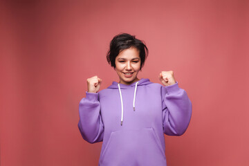 portrait of a cheerful girl with short hair, raising her fists in delight , highlighted against a pink background.