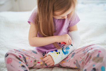 Adorable preschooler girl with a broken arm at home on the bed draws with felt-tip pens on an...