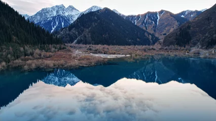 Papier Peint photo Lavable Réflexion Issyk mountain lake with mirror water at sunset. The color of the water changes before our eyes. There are trees in clear water. Snowy mountains and green hills are visible. Clouds are reflected