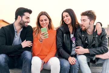 Happy group of trendy young people laughing and chatting sitting in a bench using a cellphone. Millennial diverse student friends having fun together outdoor