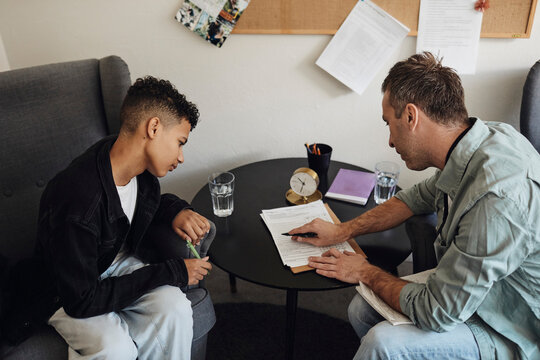 Male counselor discussing checklist with teenage student sitting at table in school office