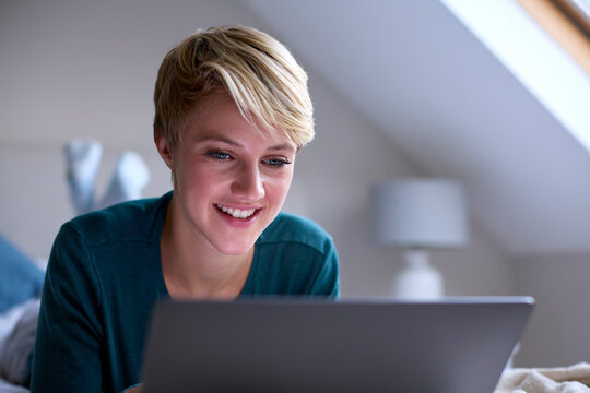 Young Smiling Woman Sitting On Bed Wearing Pyjamas At Home Working On Laptop