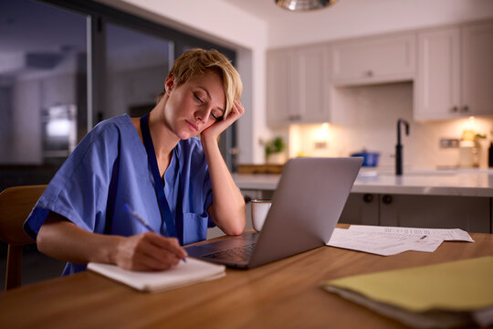 Tired Woman Wearing Medical Scrubs Working Or Studying On Laptop At Home At Night