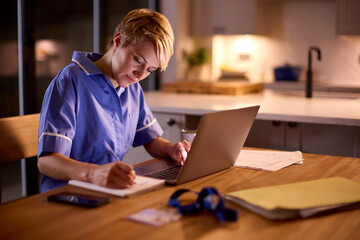 Tired Woman Wearing Medical Scrubs Working Or Studying On Laptop At Home At Night