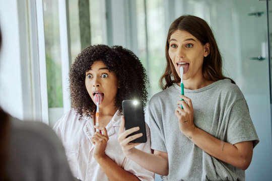 Friends, wellness and brushing teeth phone selfie with crazy face for morning hygiene routine in home. Comic, goofy and interracial friendship of women cleaning teeth with smartphone photograph.