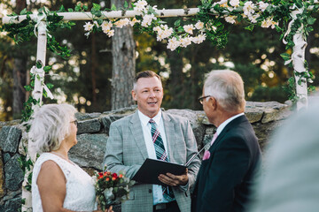 Senior couple exchanging vows while looking at minister standing against wall with flower decoration during wedding cere