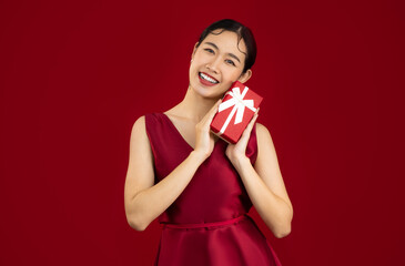 Young asian woman in red luxury dress holding and giving present red box white bow pose looking and smiling red background.